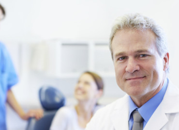 A mature dentist crossing his arms as his assistant and patient converse in the background
