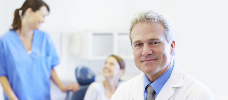 A mature dentist crossing his arms as his assistant and patient converse in the background